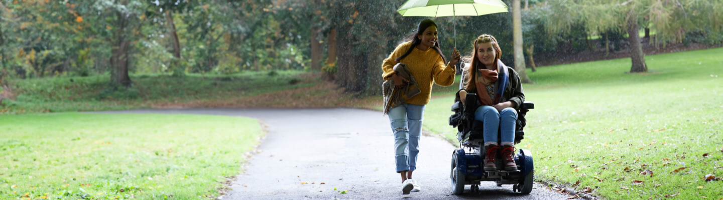2 vrouwen in de regen onder de paraplu. 1 persoon zit in een rolstoel.